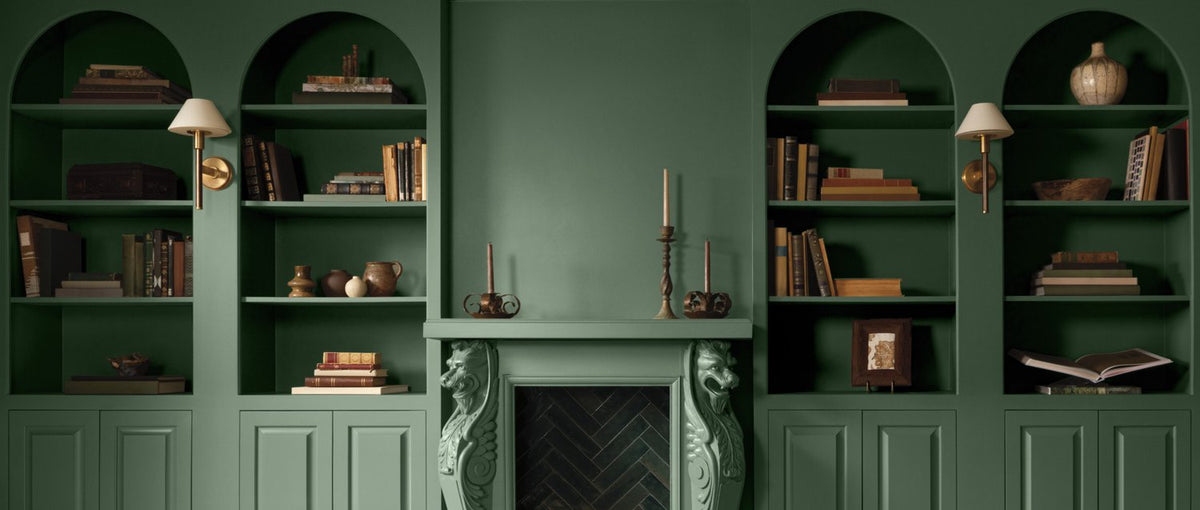 A green painted room with lower panelled walls and arched, built-in shelving topped with books and decor.